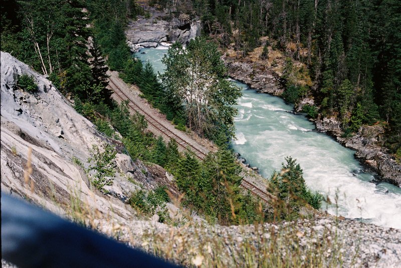 Railway tracks follow a pale blue river through a rocky forested gorge.